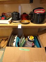 Wide photo showing assorted camping cookware on shelf and inside cardboard box including pots, pans, dish rack, dishes
