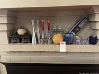 Full view of various candle holders arranged on a white shelf including metal houses, silver taper holders, red candles, blue ceramic and glass holders.