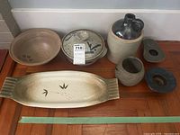 Full view of six ceramic and stoneware items arranged on wood surface including shallow bowl, lidded pot, long tray with leaf design, stoneware jug, small pot, and flower frog.