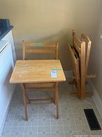 Set of 2 wood folding chairs and 2 wood TV folding trays stacked and standing against a wall, showing the chairs' backs and the tops of the trays.