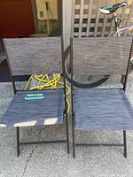 Two folding black metal chairs with mesh fabric seats and backs, placed side by side on concrete with cushions visible on the seats.