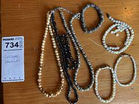 Photo of seven costume jewelry pieces including bracelets and necklaces, some pearl-style, some metallic, set on a wood table.