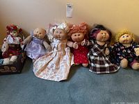 All six dolls lined up on a carpeted floor against a beige wall, showing faces and dresses in detail.