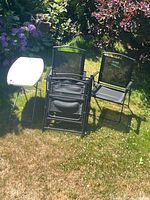 Four black folding chairs and one white folding side table arranged on grass with foliage background.