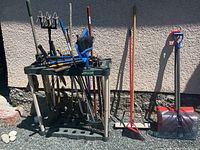 Overall view of garden tools on plastic rack leaning against wall including shovels, rakes, push broom, and hand tools