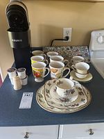 Keurig coffee maker with six coffee mugs, three tea cups and saucers, salt and pepper shakers and coffee pods displayed on countertop.