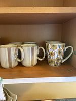 Shelf with 6 cream-colored mugs with vertical brown stripes and 1 white floral patterned mug