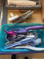 Photo of assorted kitchen utensils in a plastic container including forks, knives, peelers, and a pink handled peeler, with a handheld electric cutter in a gray case on the side.