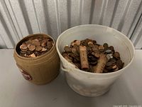 Overview photo showing two containers filled with pennies and three sealed penny rolls inside the white plastic bucket.