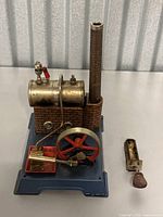 Model steam boiler with cylindrical brass tank, brick chimney, red flywheel, blue base, and separate burner accessory with red handle.