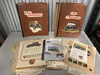 Two brown vintage albums labeled 'The Pioneers' along with a large stack of loose antique car advertisements spread out on a flat surface.