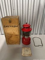 Red Coleman globe lantern with glass globe, vented top, and round red base placed next to a wooden box labeled 'LANTERN', metal handle, and instruction manual on a table.