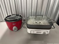 Two kitchen appliances on table: red Cuisinart rice cooker with glass lid on left and white gray Cuisinart electric grill with glass lid on right.