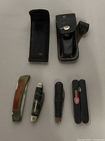 Full group image of five folding knives including two leather sheaths, laid out on white surface.