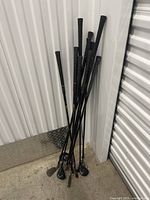 Multiple golf clubs bundled together, showing black grips and a mix of irons and woods, standing on concrete floor by a metal wall.