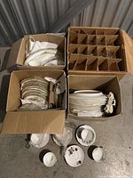 Four cardboard boxes containing stacked Mikasa china plates, bowls, and tea cups with saucers. Boxes include protective divisions for glasses or fragile items.
