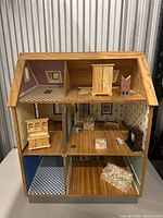 Front view of vintage wooden dollhouse showing attic, second and first floors with wallpaper and wooden floor, containing wooden furniture including wardrobe and piano