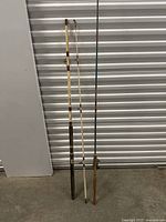 Three vintage fishing rods standing upright against a metal wall, two without reels and one with a reel attached