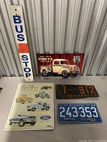 Overview of all signs and plates on table including BUS STOP sign, Ford V-8 truck sign, Montana and British Columbia plates, and Ford trucks poster