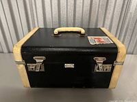 Closed black travel chest with cream edges, handle on top, and two silver latches on front. A vintage travel sticker is affixed to the top right corner.