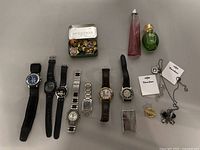 Full lot image showing eight wrist watches, two perfume bottles, metal tin with pins, jewelry, and lighter on a grey surface.