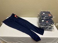 One pair navy blue pants laid flat beside stack of nine packaged pairs