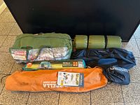 Six packed items including Coleman tent bag, rolled green pad, black bags, orange Sport-Brella chair bag