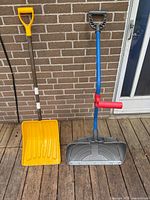 Both snow shovels standing against brick wall showing blades, shafts, grips and auxiliary handle