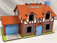 Front and side view of the vintage Fisher Price Little People brown Tudor style play house with garage attached. Red roof with Fisher Price logo on top and functional blue front door and garage door.