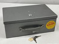 Image of closed rectangular gray metal strongbox with carrying handle and fireproof certification sticker on front, accompanied by two keys placed in front.