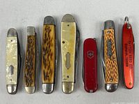 Seven vintage pocket knives laid flat showing front handles in various materials: pearl-like, imitation stag, mottled bone, red plastic with logo, and advertising.