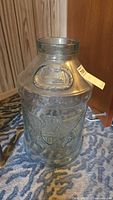 Clear five-gallon glass jug standing on a patterned rug next to wood paneling. The jug has a tag attached and wide mouth opening.
