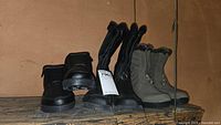 Full lot of six men's and ladies' boots on wooden shelf showing black snow boots, mid-calf lace-up snow boots, and greenish-grey boots.