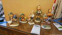 Front view of all six Goebel Hummel figurines placed in a row on a wooden surface with a beige background. Figurines show children with animals and various objects.