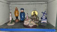 Shelf display of seashore themed decor items including lighthouse snow globe, captain figurines, Lenox lighthouse, and windmill music box.
