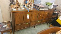 Front view of mid-century modern sideboard showing 3 cabinet doors, 5 drawers, wood grain finish, brass hardware, with various decorative items on top.
