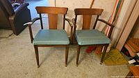 Front view of two matching mid century modern wood dining arm chairs with light blue upholstered seats, tapered legs and brass feet caps.