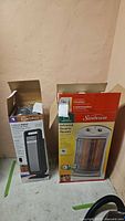 Two boxed space heaters side by side against wall: Lasko Digital Ceramic Tower Heater and Sunbeam Infrared Quartz Heater.