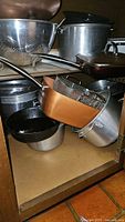 Various pots and pans including stainless colander, enamelware pot, aluminum pot, copper pan with basket, cast iron skillet in cabinet.