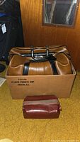 Vintage brown leather saddle bag with black vertical straps and handles packed inside an Atlantic Products Corp. branded cardboard box on a yellow carpet. A small maroon leather travel case is placed in front of the box.
