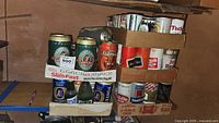 Wide shot showing multiple cardboard trays holding vintage beer cans and some glass bottles on basement shelf.