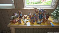 Full view of all 17 ceramic nativity figurines displayed on a yellow felt base by a window, including human characters, camels, and animals.