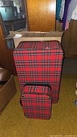 Photo of the two red plaid luggage pieces, one large upright case and one smaller carry-on case in front of original cardboard box showing signs of wear.