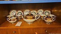 Tea set arranged with six cups and saucers, lidded sugar bowl, creamer, and lidded teapot, all on a wooden shelf, showing gold trim and overall design.