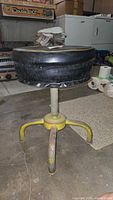 Side view of vintage Ajusto metal stool showing round padded black seat and yellow metal leg base with rust and wear.
