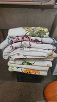 Stack of vintage large towels with various printed patterns folded on a crate in basement setting.
