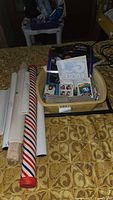 Photo showing beige plastic tub containing trading cards and loose printed materials, with rolls of wrapping paper with red, white, and blue stripes beside it on a patterned yellow surface.