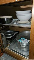 Shelf with white ceramic bowls stacked, metal mixing bowls, and aluminum baking sheets including rectangular pie tins