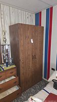 Front view of double door wardrobe cabinet with woodgrain finish next to a dresser and colorful wall.