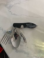 Folded 4-in-1 folding camping utensil showing fork, spoon, cutting utensil, and bottle opener with black handle labeled 'Castle Weld Shop'